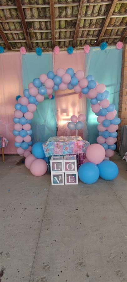 Arco di palloncini rosa e azzurri sotto un portico, con teli chiari e le lettere LOVE sul tavolo