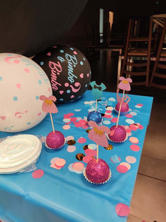 Tavolo con tovaglia azzurra, cupcake con topper rosa e azzurri, coriandoli tondi e palloncini